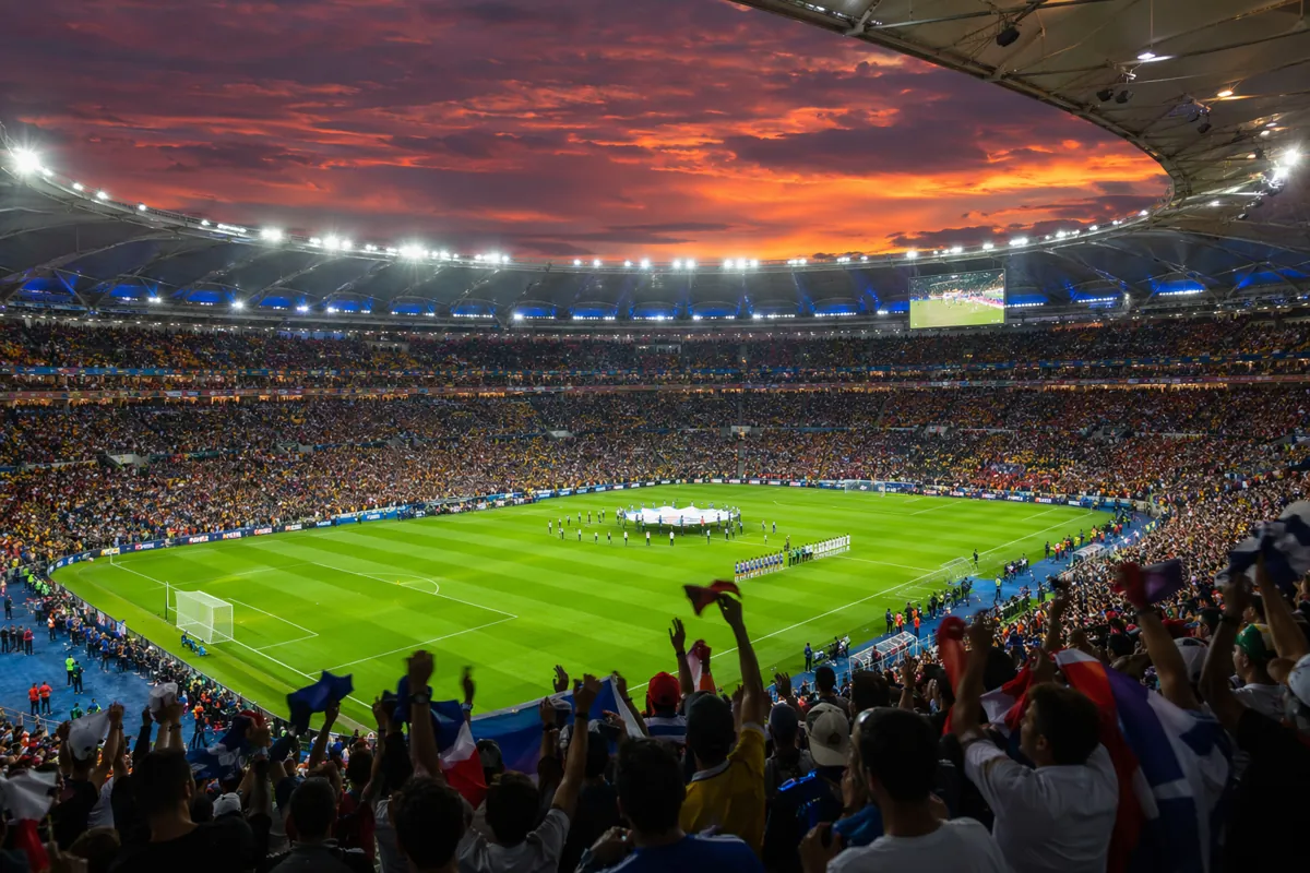 Wide shot of a professional soccer stadium at dusk, bright floodlights illuminating the gr